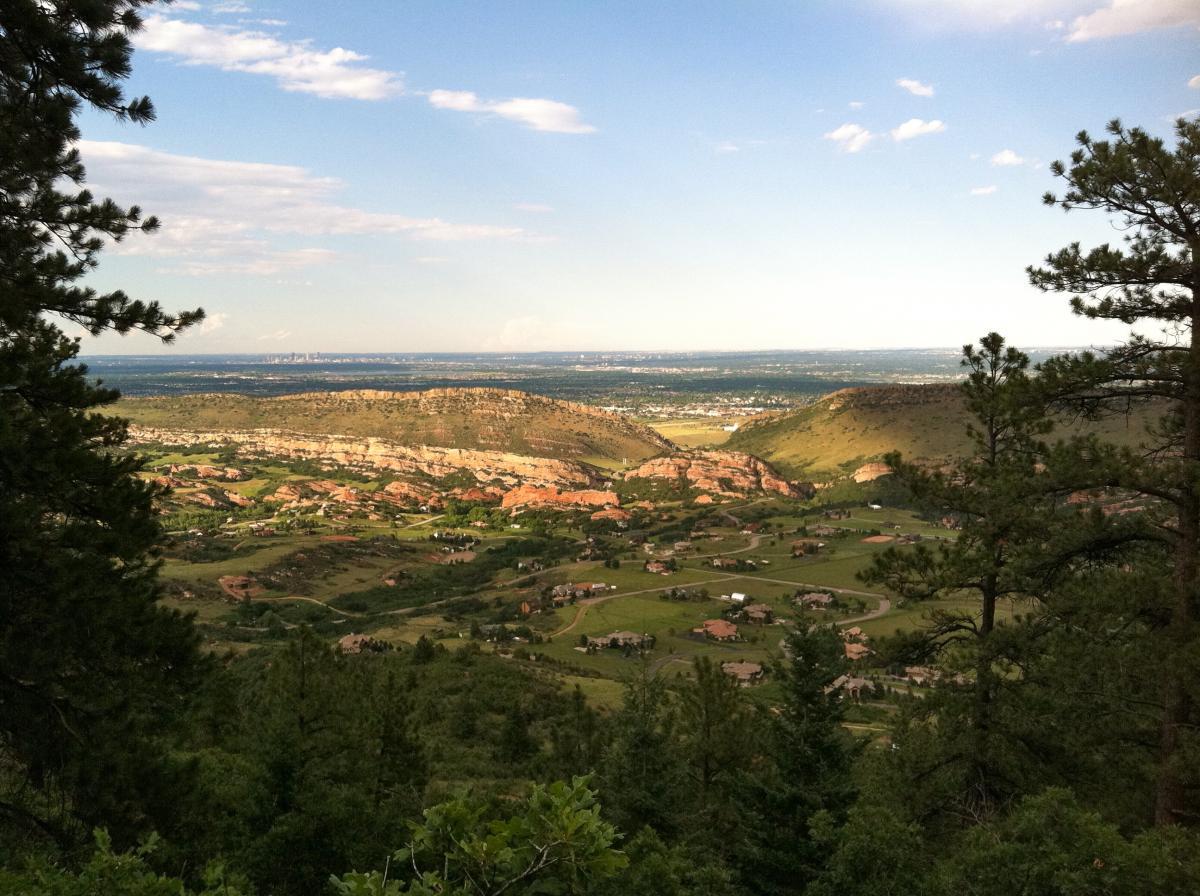 A scenic view of rolling green hills and rocky outcrops under a blue sky, with scattered residential areas visible among the lush landscape. Pine trees frame the foreground, offering a glimpse of a distant city skyline on the horizon. Deer Creek Canyon mountain bike trail.