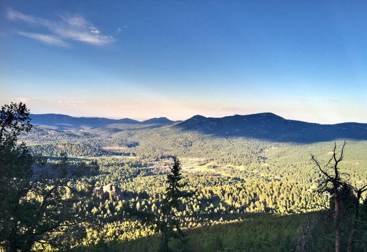 A panoramic view of a green mountain valley surrounded by rolling hills, under a clear blue sky. The landscape features dense forests of pine trees, rocky outcrops, and a small town visible in the distance. The scene is bathed in natural light, highlighting the vibrant greens of the vegetation. 3 Sisters / Alderfer mountain bike trail.
