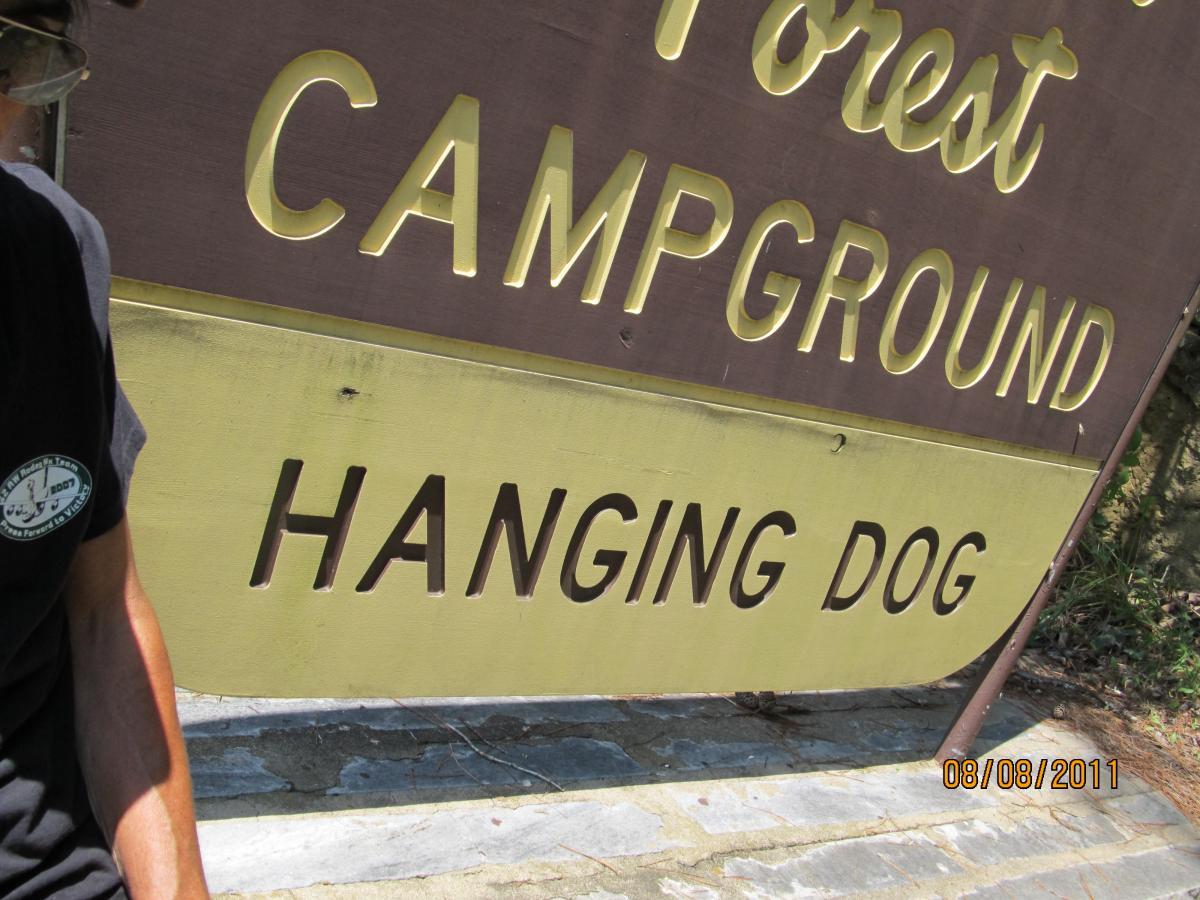 A partially visible person in a black shirt next to a campground sign that reads "HANGING DOG" with the words "FOREST CAMPGROUND" above it. The background shows a sunny, outdoor setting. Hanging Dog mountain bike trail.