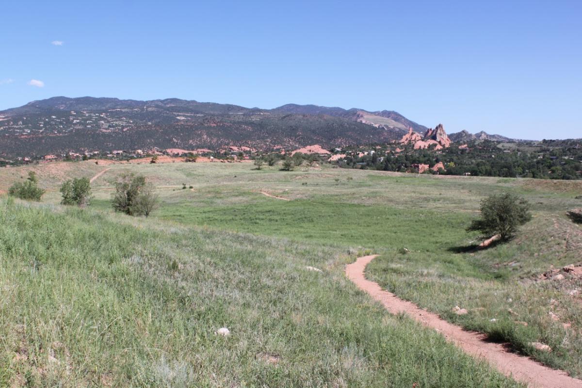 A scenic landscape showcasing rolling green fields, a dirt path winding through the foreground, and mountains in the background under a clear blue sky. The area features a mix of trees and rocky formations, suggesting a natural park or outdoor recreation area. Red Rock Canyon mountain bike trail.