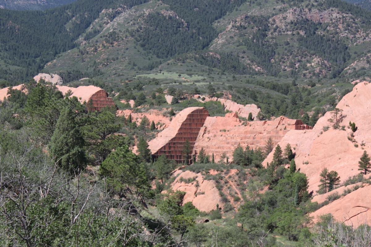 A scenic view of a rocky landscape featuring reddish sandstone formations and green vegetation, with hills and mountains in the background under a clear blue sky. Red Rock Canyon mountain bike trail.