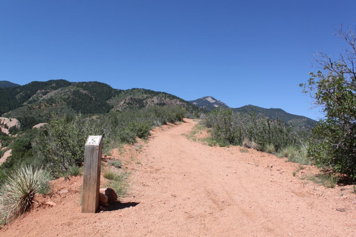 A sunny hiking trail winding through a mountainous area, with a wooden trail marker on the left side. The path is sandy and surrounded by shrubs and low plants, leading toward the distant mountains under a clear blue sky. Red Rock Canyon mountain bike trail.