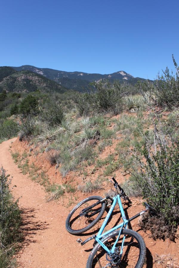 A mountain bike lying on a dirt trail surrounded by shrubs and grass, with a backdrop of rolling hills and a clear blue sky. Red Rock Canyon mountain bike trail.