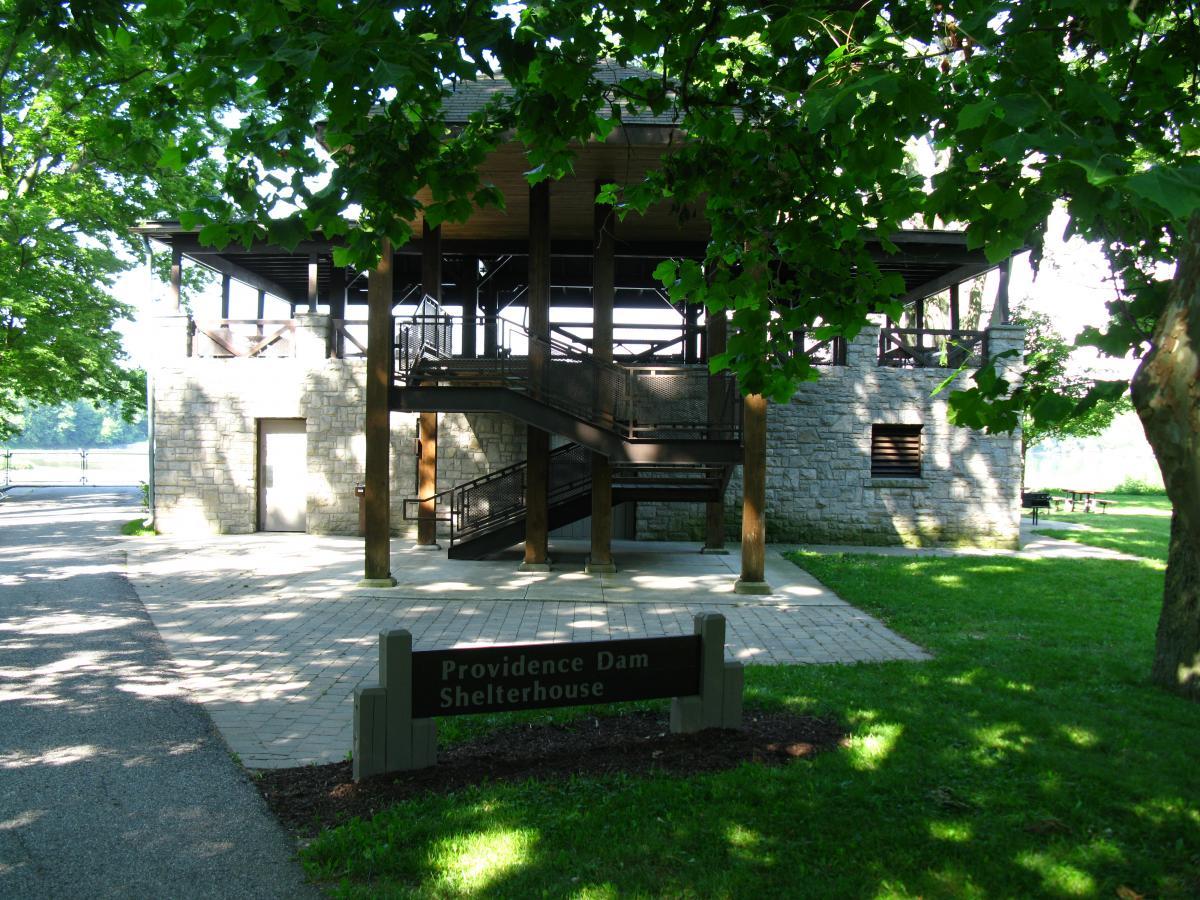A two-story shelterhouse made of stone and wood, located near Providence Dam. The structure features a stairway leading to the upper level and is surrounded by green grass and trees. A sign at the front identifies the building as "Providence Dam Shelterhouse." A gravel walkway leads up to the entrance, with picnic tables visible in the background. Farnsworth mountain bike trail.