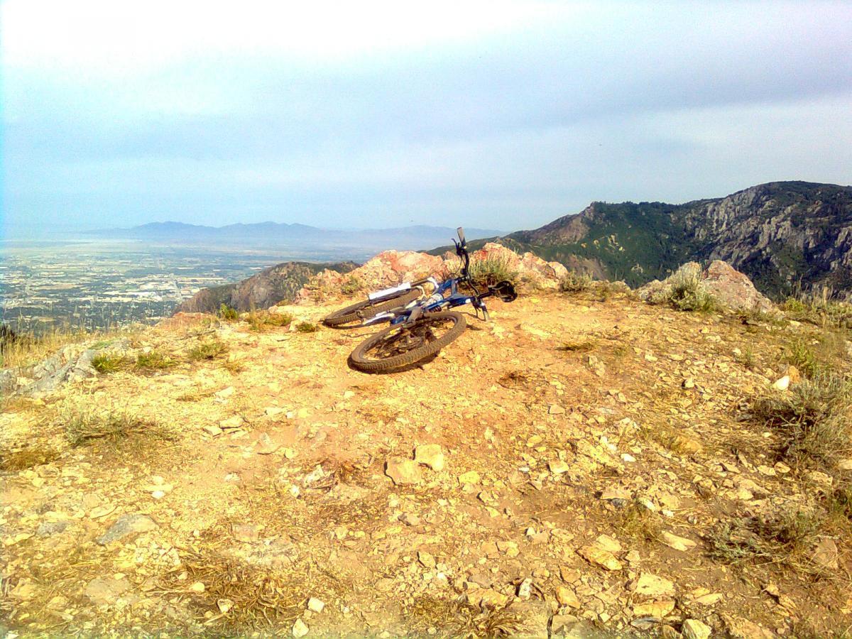 A mountain bike resting on rocky terrain at the summit, with a panoramic view of a valley and distant mountains under a partly cloudy sky. Sardine Peak Loop mountain bike trail.
