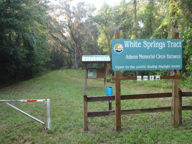 Sign at the entrance to the White Springs Tract, showing a path surrounded by dense greenery, with a wooden gate and an interpretive information board. The sign indicates that the area is open to the public during daylight hours. Bridge to Bridge mountain bike trail.