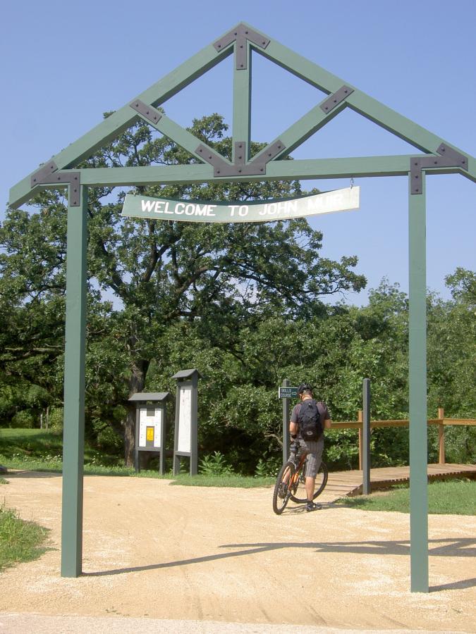 A cyclist stands with a mountain bike near the entrance of a park, marked by a green archway that reads "Welcome to John Muir." The scene features a dirt path leading into the park, surrounded by greenery and trees, with informational signs visible nearby. John Muir Trails mountain bike trail.
