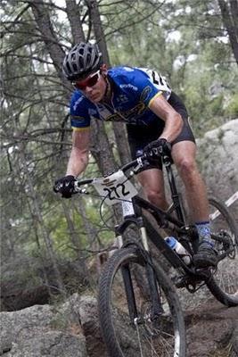 A cyclist in a blue and yellow jersey navigates rocky terrain on a mountain bike, wearing a helmet and sunglasses. The background features trees and natural scenery, indicating an outdoor biking trail. The cyclist's number is visible on the front of the jersey. Palmer Park mountain bike trail.
