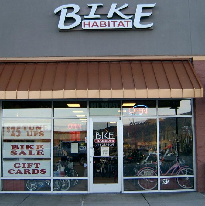 Image of the front entrance of a bike shop called "Bike Habitat." The store features a large sign with the name prominently displayed, alongside a red "OPEN" sign in the window. The storefront includes promotional messages for services like "$25 Tune Ups," "Bike Sale," and "Gift Cards," with bicycles visible through the windows. The building has a brown awning and a modern design.