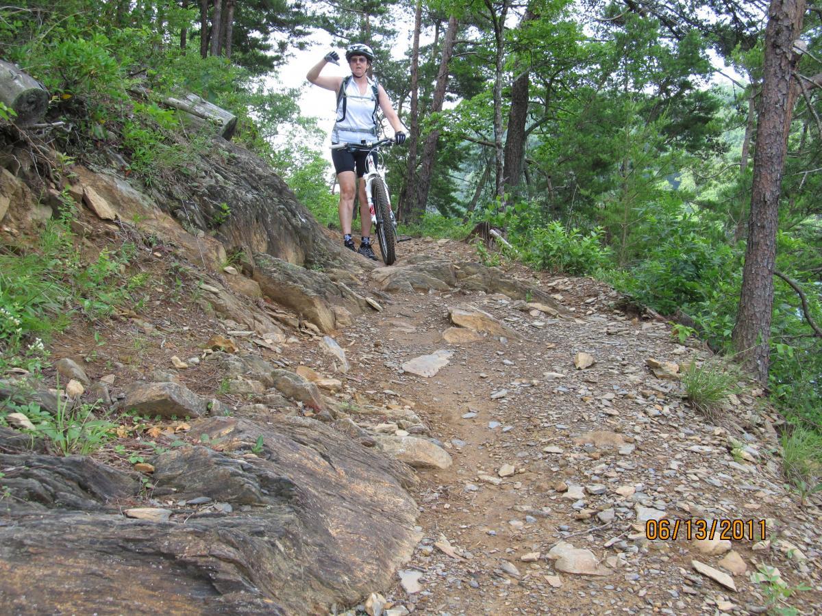 A mountain biker pauses on a rocky trail surrounded by trees. The cyclist, wearing a helmet and a backpack, raises their hand in a gesture, possibly to wave or signal. The path is uneven with large stones and dirt, indicating a rugged outdoor environment. Tsali Left Loop mountain bike trail.