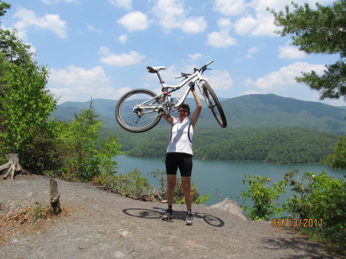 A person stands on a rocky outcrop by a lake, raising a mountain bike above their head. The background features lush green mountains and a clear blue sky with scattered clouds. The scene captures a sense of celebration and adventure. Tsali Left Loop mountain bike trail.