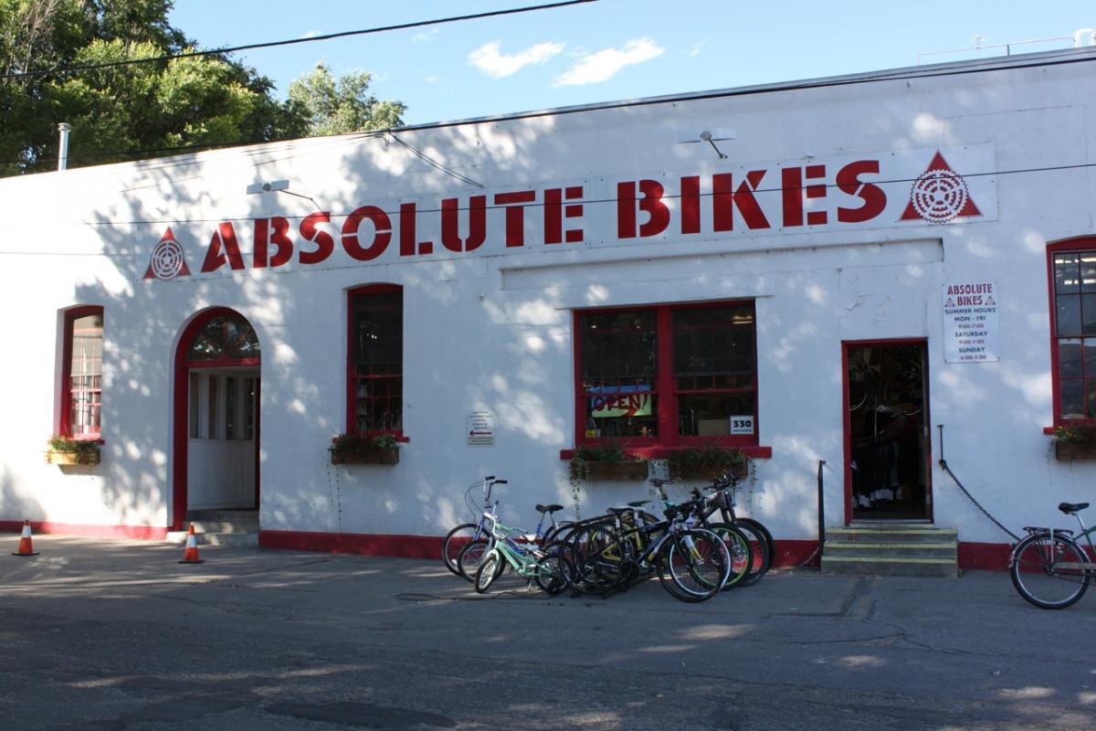 Image of a bike shop exterior featuring the name "Absolute Bikes" prominently displayed in bold red letters. The building has white walls and red accents, with several bicycles parked in front. Windows are open with red trim, and a sign detailing store hours is visible next to the entrance. The setting includes trees in the background and a clear blue sky.
