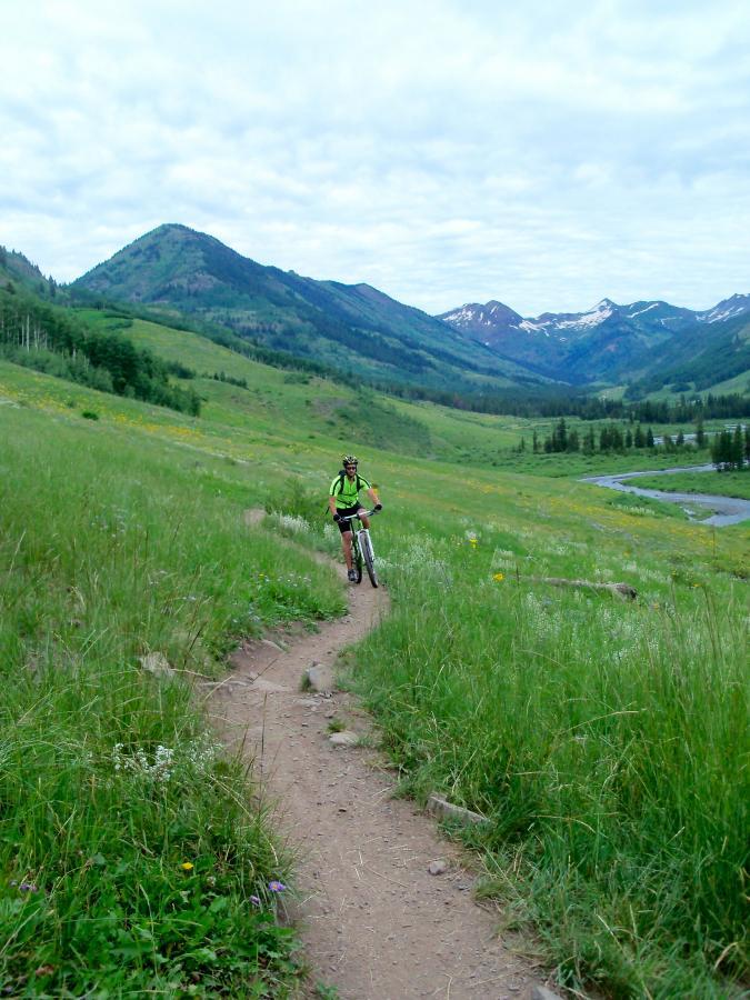 A mountain biker riding on a winding dirt trail through lush green grass, surrounded by mountains and a cloudy sky. Wildflowers dot the landscape, and a river can be seen in the background. Upper Lower Loop mountain bike trail.