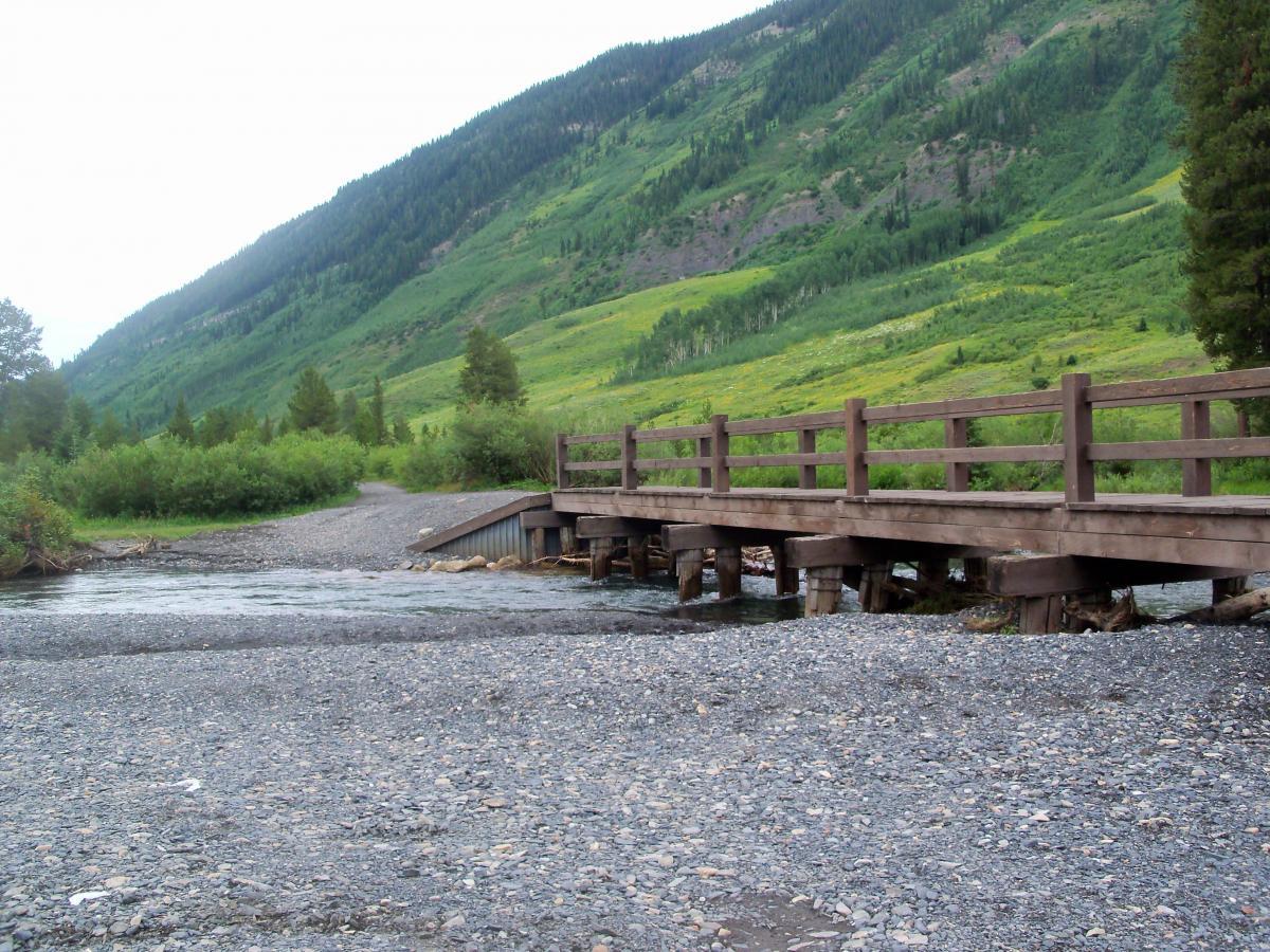 A wooden bridge spans a shallow stream, surrounded by lush green hills and trees. The landscape features a rocky shore and a winding path leading into the distance. Lower Loop mountain bike trail.