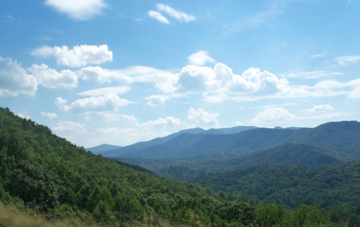 A scenic view of rolling mountains under a blue sky adorned with fluffy white clouds, surrounded by dense greenery in the foreground. Brush Creek mountain bike trail.