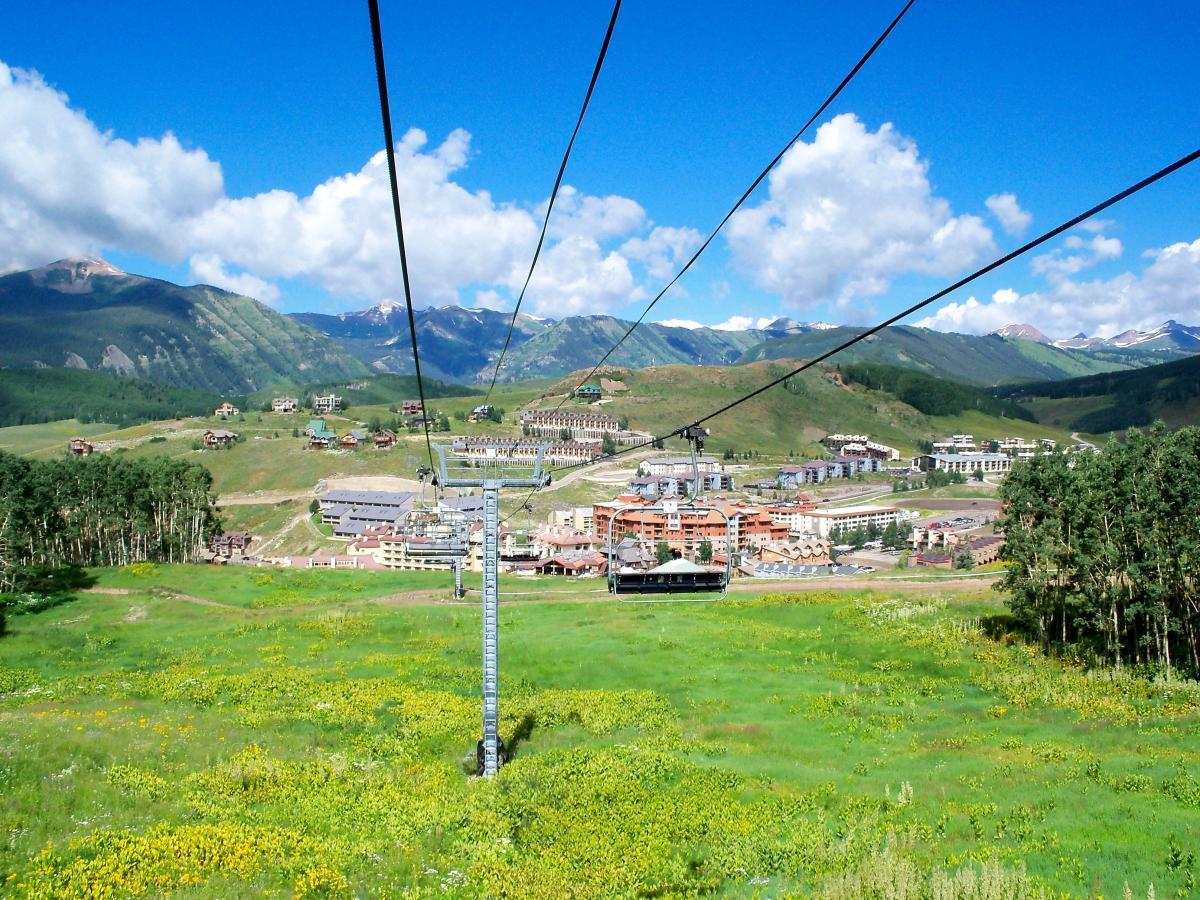 A scenic view from a ski lift showing a vibrant summer landscape with green fields and wildflowers. In the background, the mountains rise majestically under a blue sky adorned with fluffy white clouds. Below, a resort community is visible, featuring buildings nestled in the valley and surrounded by lush trees. Evolution Bike Park at Crested Butte Mountain Resort mountain bike trail.