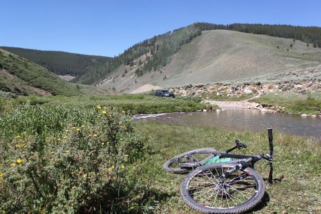 A mountain bike lying on its side in a grassy area, with a stream flowing nearby. In the background, rolling hills covered in lush greenery and trees can be seen under a clear blue sky. A vehicle is parked in the distance along a dirt road. Doctor Park mountain bike trail.