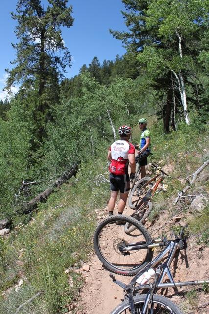 Two mountain bikers pause on a dirt trail surrounded by green trees and vegetation. One biker is wearing a red and white jersey with a black helmet, and the other is in a green and blue jersey with a green helmet. Several bicycles are leaning against the trail, with the front wheel of one bike prominently in the foreground. Doctor Park mountain bike trail.