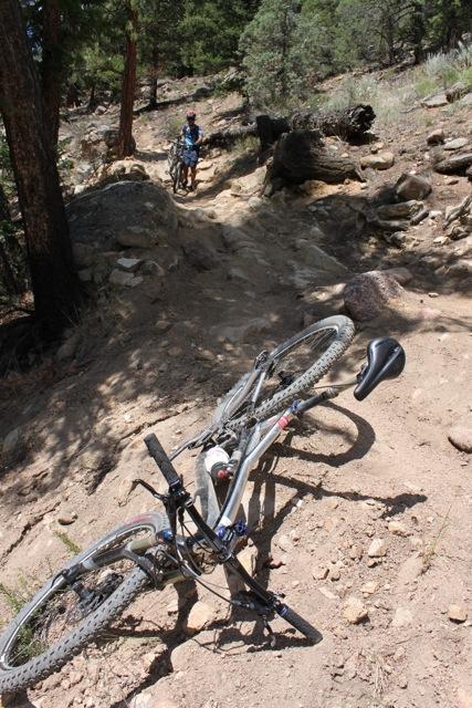 A mountain bike lying on its side on a rocky, dirt trail surrounded by trees, with a hiker in the background walking along the path. Doctor Park mountain bike trail.
