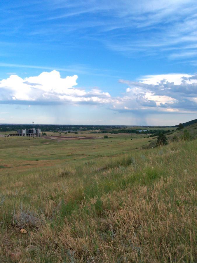 A scenic view of an open landscape featuring rolling hills, grassy fields, and a partly cloudy sky. In the foreground, wild grasses and small plants are visible, while the background shows distant hills under a blue sky with white clouds. A structure can be seen to the left, adding context to the natural surroundings. Foothills Trail mountain bike trail.
