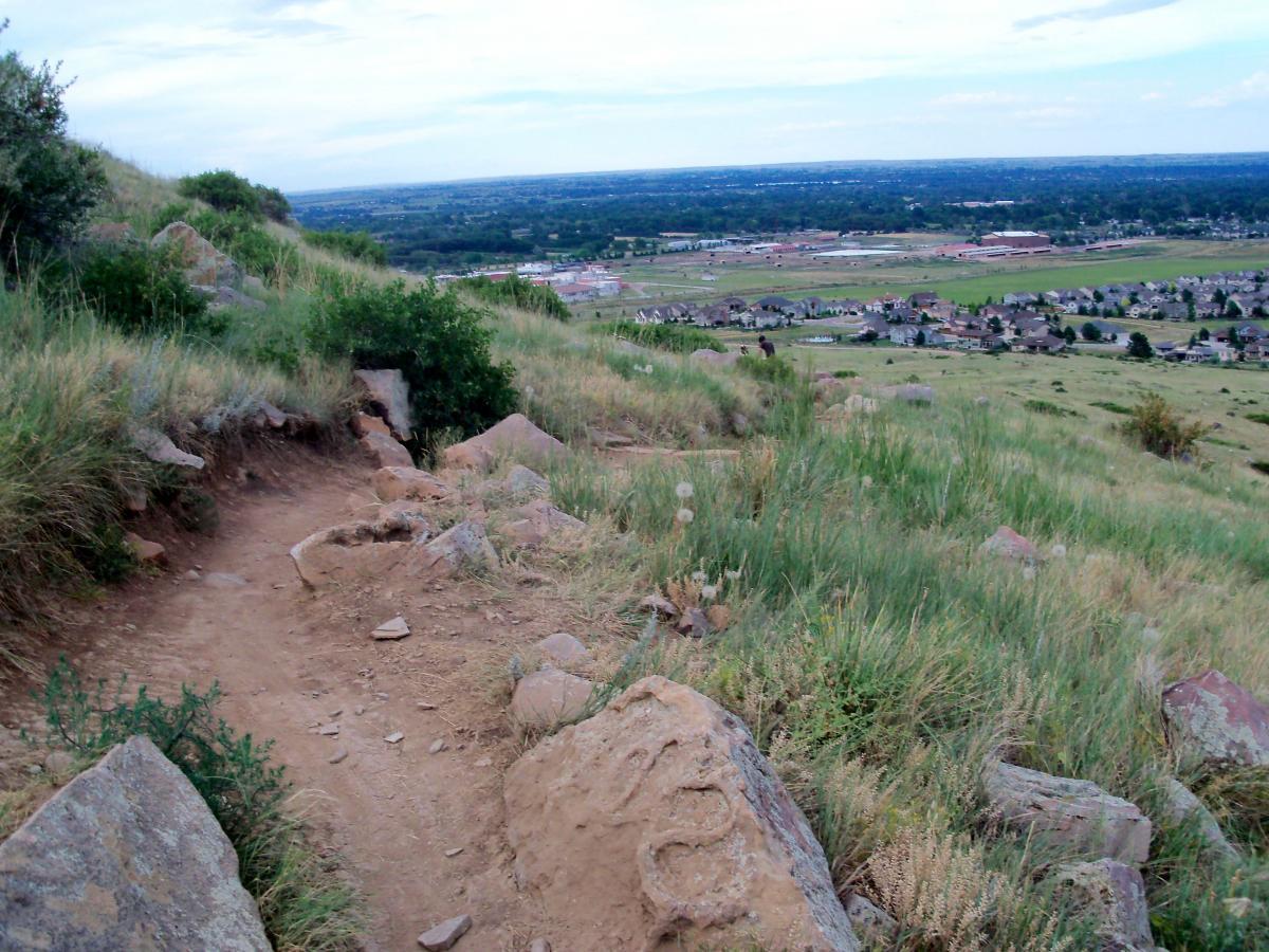 A winding dirt trail surrounded by green grass and rocky terrain, leading down a hillside with a view of a distant valley and residential area below. The sky is partly cloudy, and the landscape features scattered rocks and vegetation. Foothills Trail mountain bike trail.