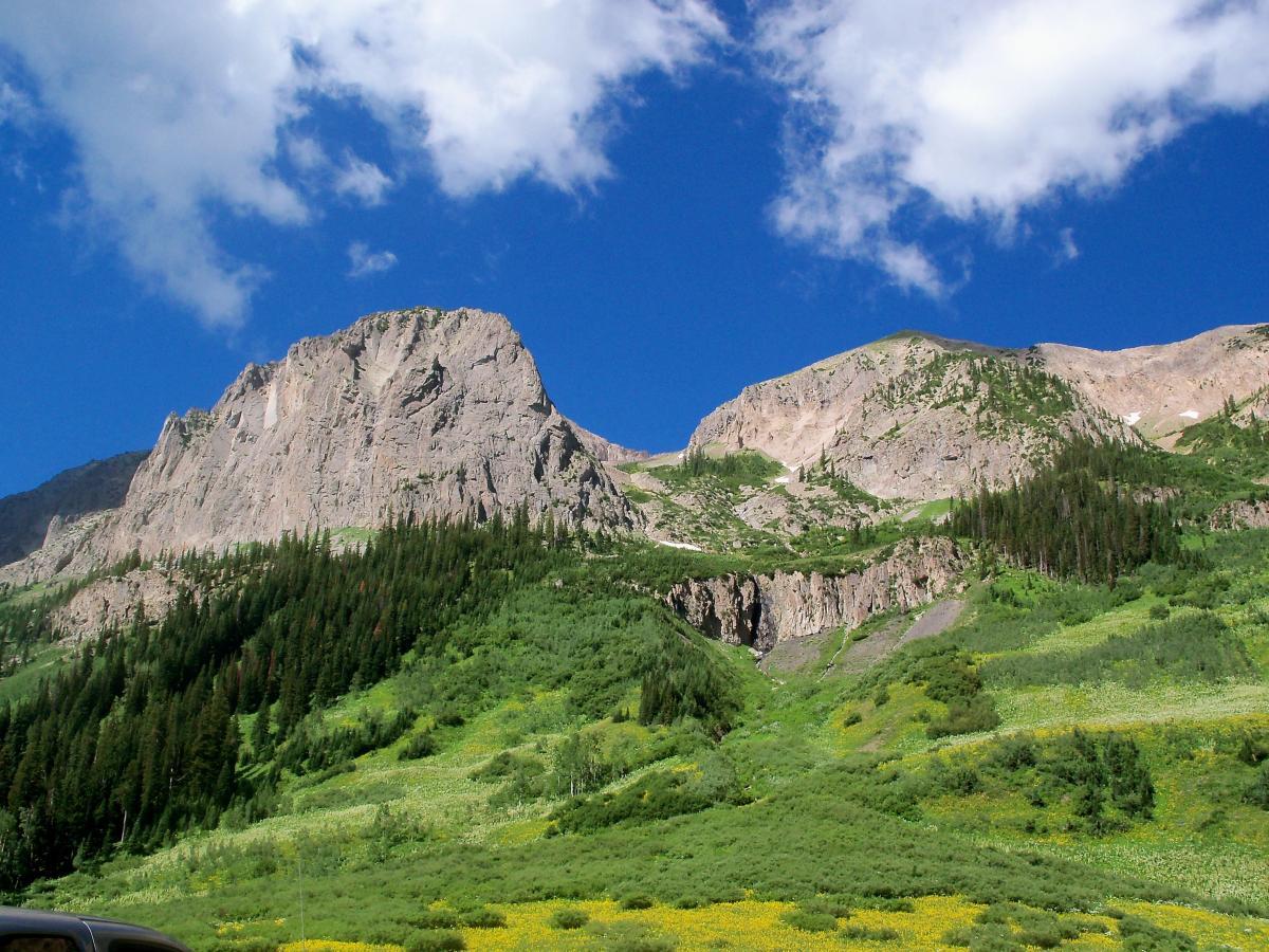 A panoramic view of rugged mountain peaks under a bright blue sky, with scattered fluffy clouds. Lush green trees and flowering meadows cover the lower slopes, showcasing a vibrant natural landscape. A parked vehicle is visible in the foreground, adding a sense of scale to the majestic scenery. Trail 401 mountain bike trail.