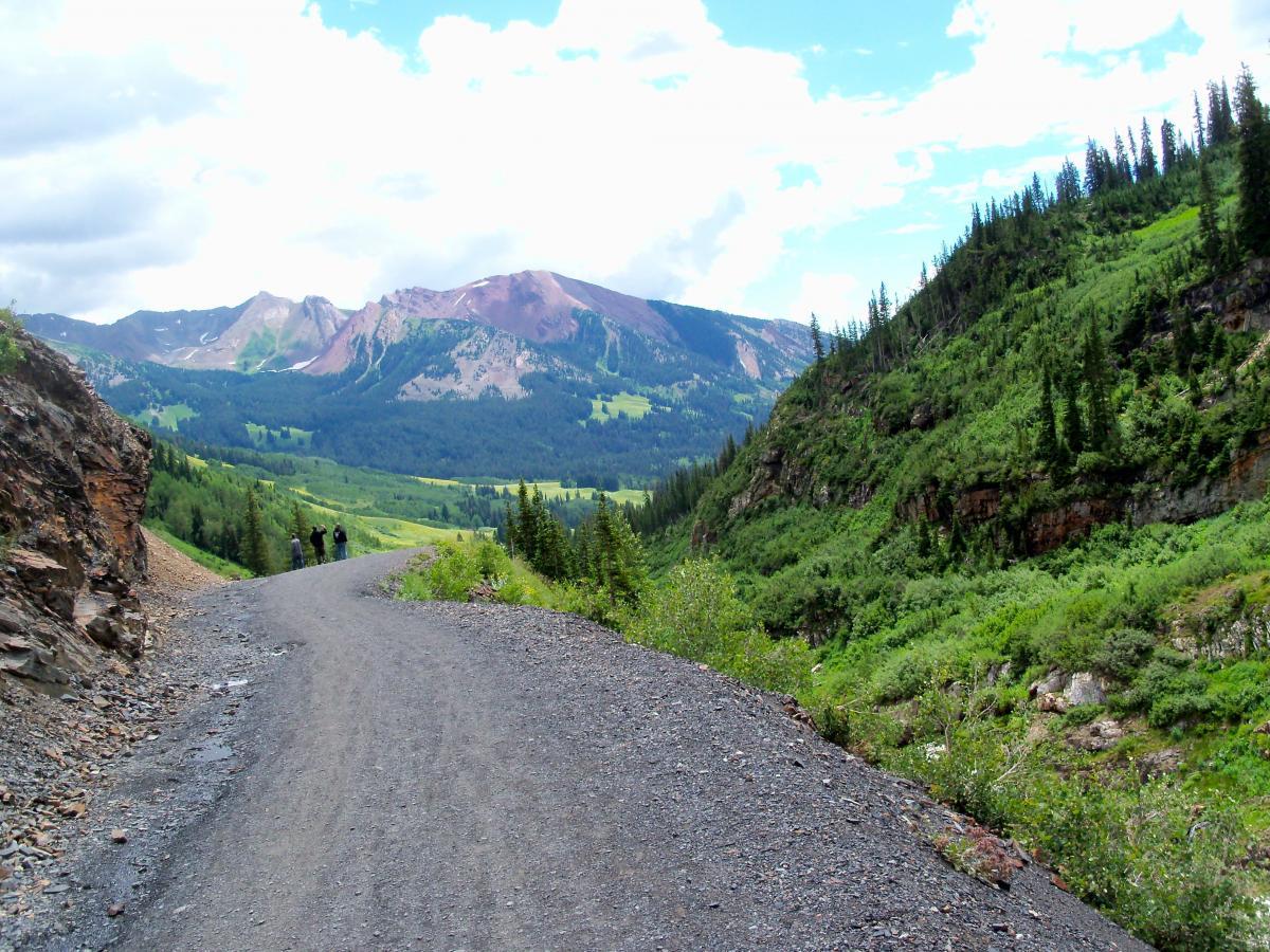 A scenic trail winding through a lush mountainous landscape, with a gravel path leading into the distance. In the background, green hills and rugged mountains are visible under a partly cloudy sky. Two hikers can be seen walking along the trail, surrounded by vibrant greenery and trees on either side. Trail 401 mountain bike trail.