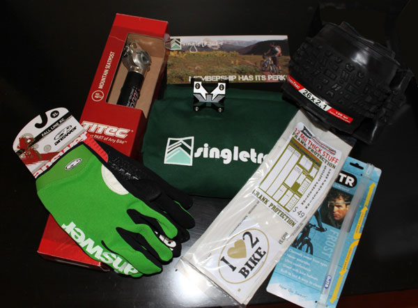 A flat lay arrangement of cycling accessories including a green pair of gloves, various bike components in their packaging, a t-shirt with the "singletrack" logo, a membership card, and stickers that read "I Love Bike." The items are displayed on a dark surface, showcasing a variety of gear for biking enthusiasts.