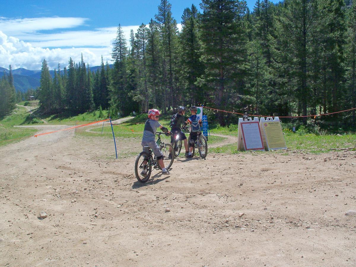 Two children in helmets are ready to ride their bicycles on a dirt path surrounded by tall trees and mountains in the background. A sign indicates trail information, and an orange barrier tape marks the path. The sky is bright with scattered clouds. Evolution Bike Park at Crested Butte Mountain Resort mountain bike trail.