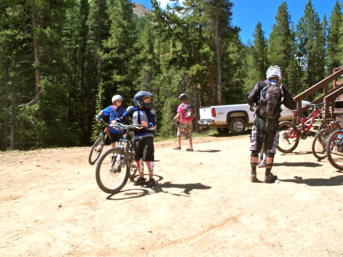 A group of young mountain bikers, wearing helmets and protective gear, stand on a dirt path surrounded by trees. Some are positioned near their bikes, while others are looking around. A pickup truck is parked nearby, and additional bicycles can be seen at a bike rack. The scene captures a sunny day in a wooded area, suggesting a fun outdoor adventure. Evolution Bike Park at Crested Butte Mountain Resort mountain bike trail.