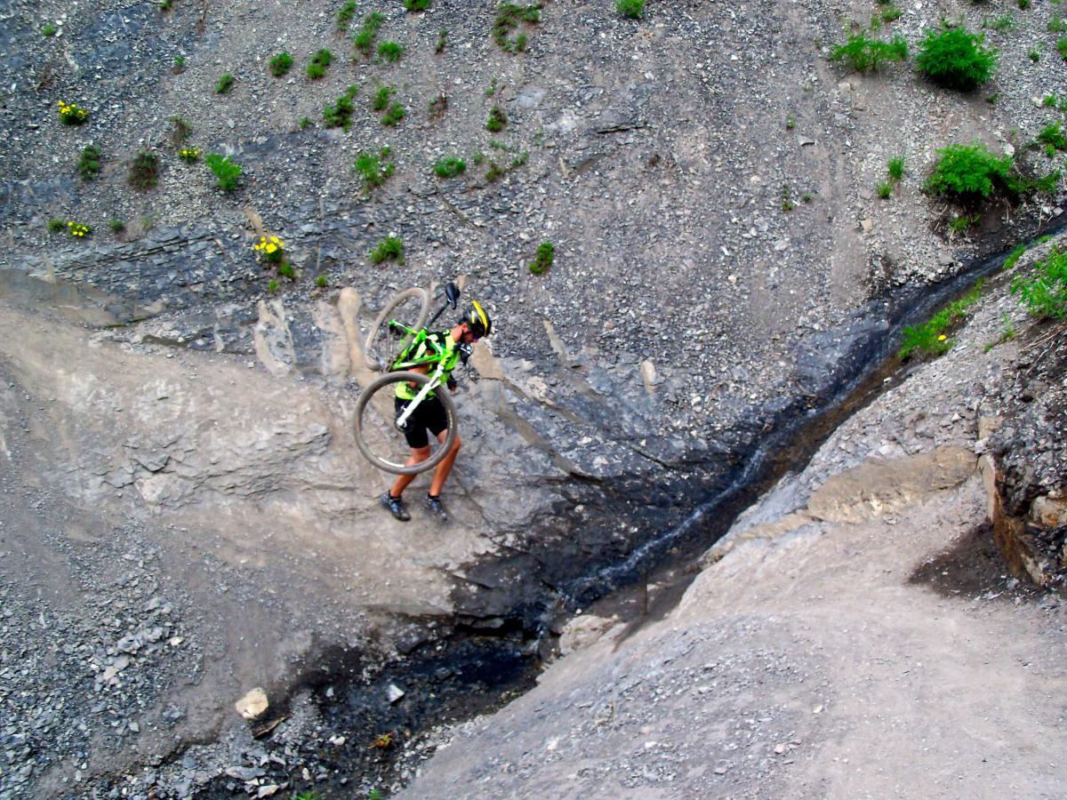A cyclist wearing a green and black outfit is carrying a mountain bike while navigating a rocky terrain. The background features sparse vegetation and rocky surfaces, indicating a rugged outdoor environment. Trail 401 mountain bike trail.