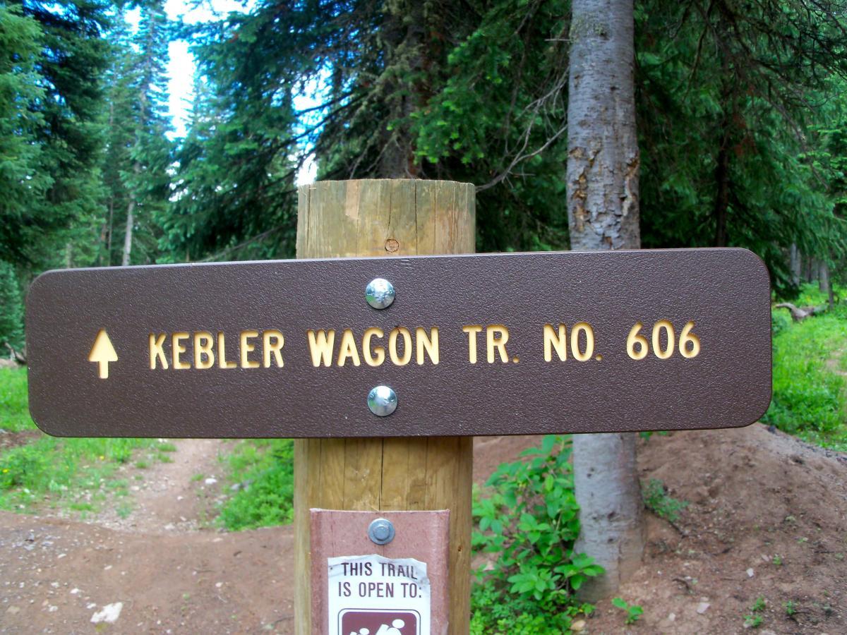 Trail sign indicating "Kebler Wagon Tr. No. 606," with an arrow pointing upward. The sign is mounted on a wooden post in a forested area surrounded by tall trees and greenery. A smaller sign below states, "This trail is open to [specific use]." Kebler Wagon Trail / #606 mountain bike trail.