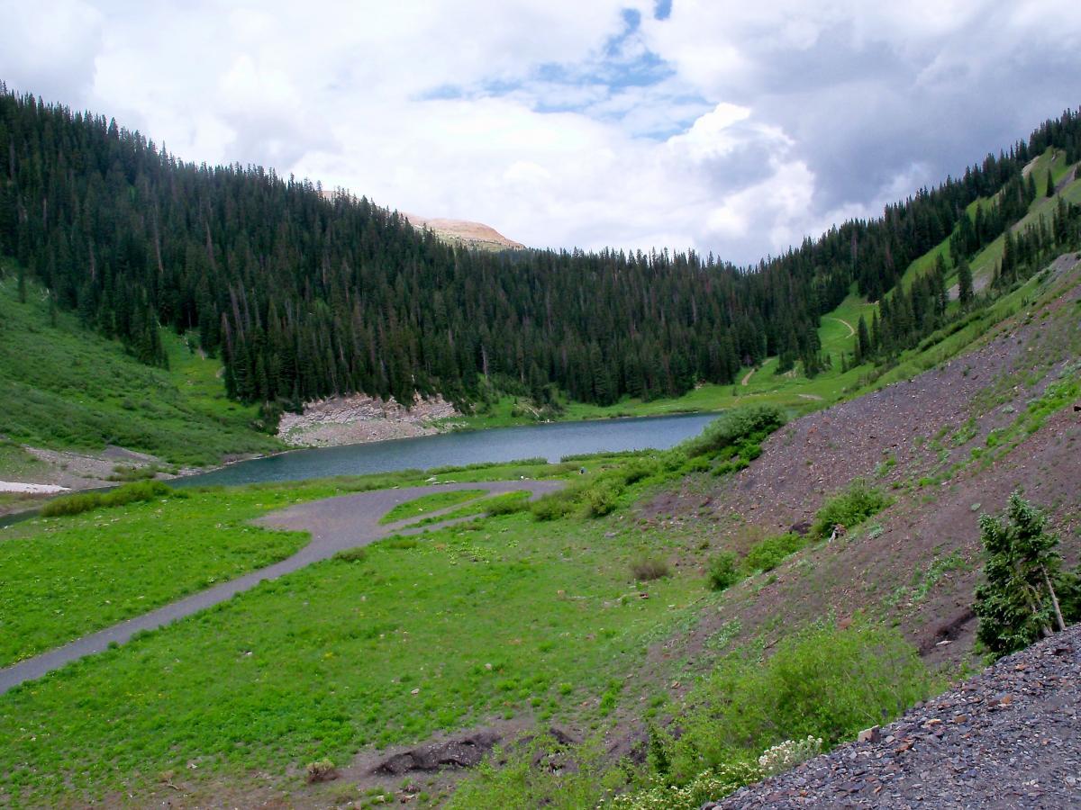 A serene mountain landscape featuring a tranquil lake surrounded by lush greenery and dense pine forests. The sky is partly cloudy, with patches of blue peeking through. Gentle slopes of grass and rocks lead to the water's edge, inviting a sense of peace and natural beauty. Trail 401 mountain bike trail.