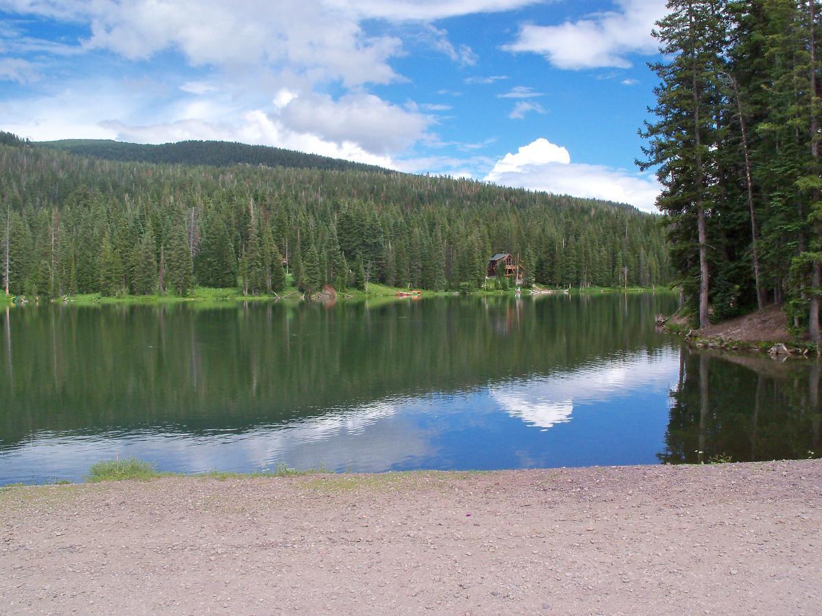 A serene lake surrounded by dense evergreen trees, reflecting the blue sky and fluffy white clouds above. The water is calm, with a sandy shore visible in the foreground. A cabin can be seen in the background, nestled among the trees. Dyke mountain bike trail.