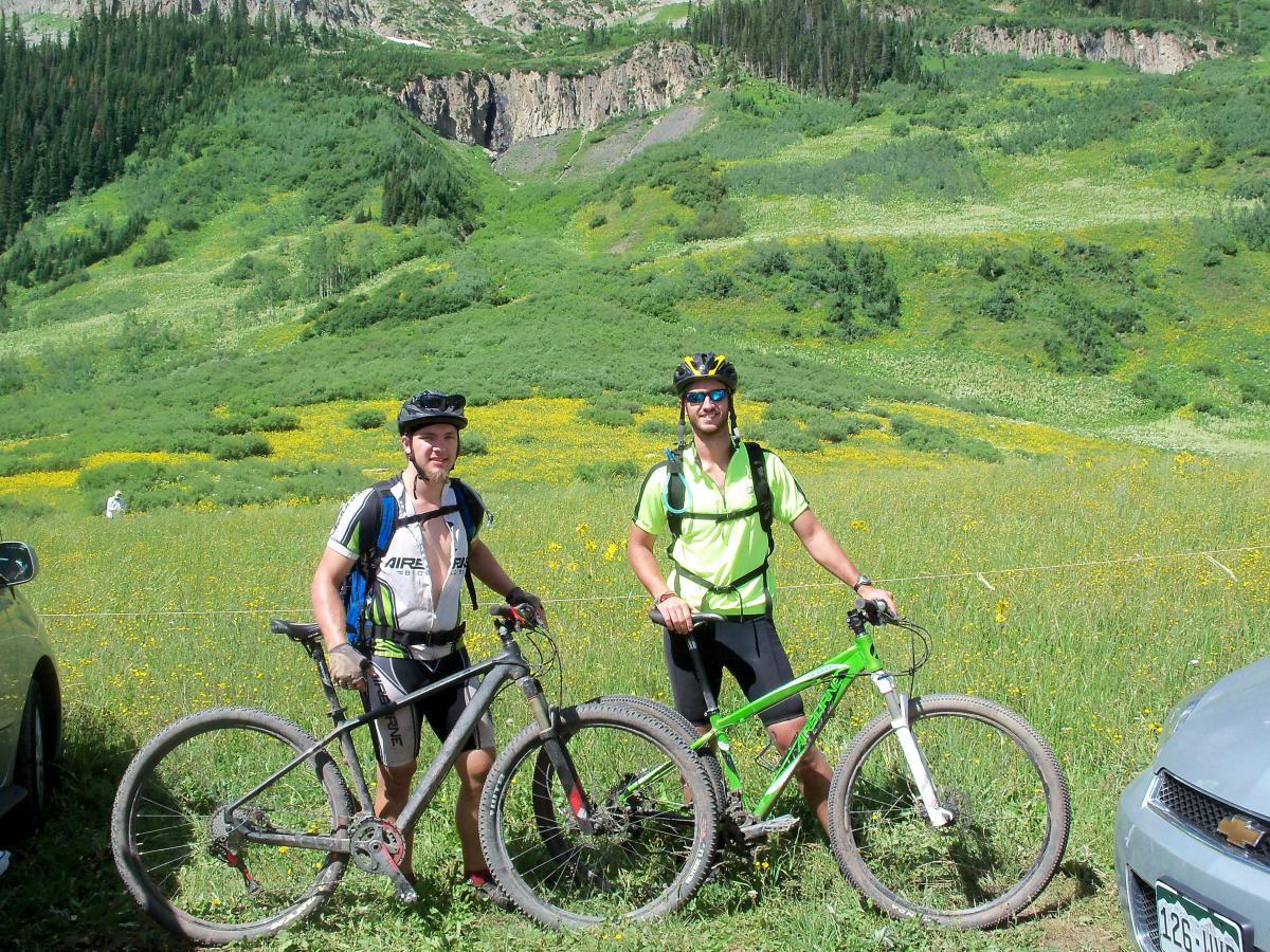 Two mountain bikers stand beside their bikes in a green, flower-filled meadow with a forested hillside in the background. One rider is wearing a black helmet and a colorful cycling jersey, while the other wears a bright green shirt and sunglasses. Both bikes are suited for off-road riding. Trail 401 mountain bike trail.