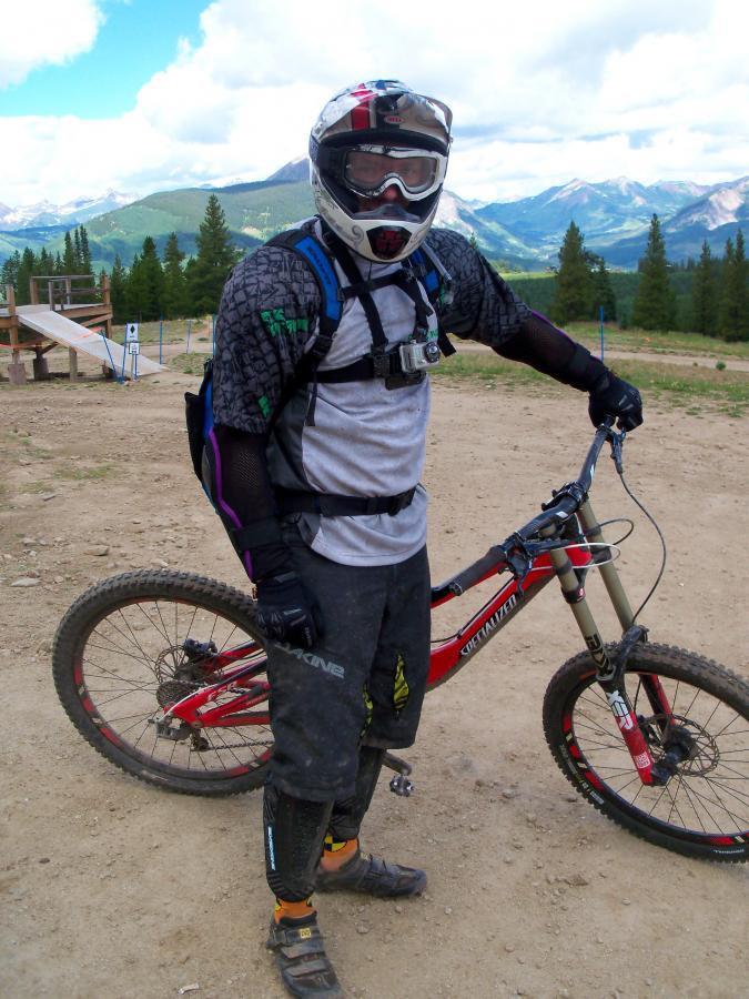 A mountain biker wearing a helmet, goggles, and protective gear, standing next to a red bike on a dirt trail with a mountainous landscape in the background. Evolution Bike Park at Crested Butte Mountain Resort mountain bike trail.