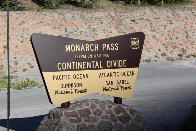 A sign marking Monarch Pass, featuring the text "MONARCH PASS" at the top, indicating an elevation of 11,312 feet. Below, the sign highlights the Continental Divide, with directions toward the Pacific Ocean and Atlantic Ocean, along with references to nearby Gunnison National Forest and San Isabel National Forest. The background shows a rocky hillside and greenery. Monarch Crest Trail mountain bike trail.