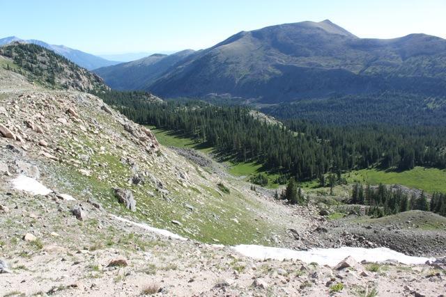A panoramic view of a mountainous landscape featuring rocky slopes, lush green valleys, and distant peaks under a clear blue sky. The scene captures the natural beauty of the terrain, with scattered trees and patches of snow visible in the foreground. Monarch Crest Trail mountain bike trail.