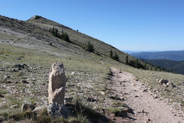 A rocky path leading uphill through a grassy landscape, with a prominent stone marker in the foreground. Pine trees line the slope, and the clear blue sky stretches above the distant mountains. Monarch Crest Trail mountain bike trail.