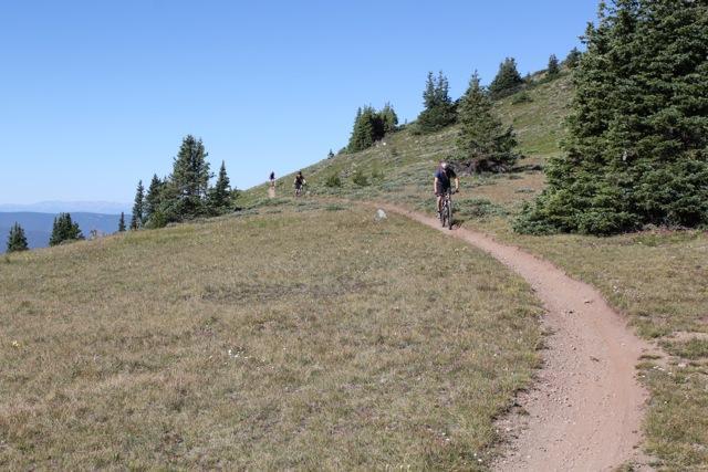 A wide, grassy trail in a mountainous area, with a cyclist riding along a dirt path. In the background, additional cyclists can be seen on the trail. The landscape features patches of trees and a clear blue sky. Monarch Crest Trail mountain bike trail.