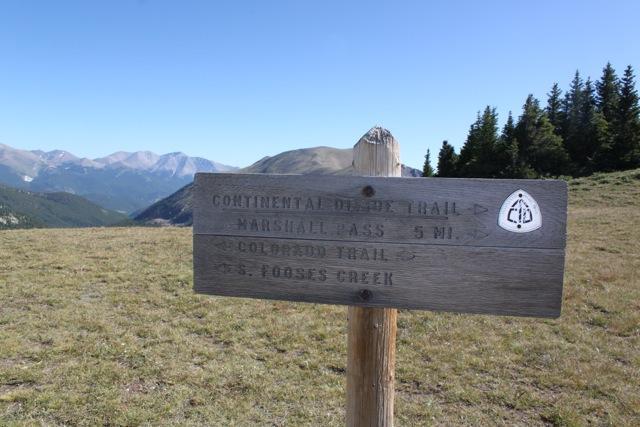 A wooden trail sign indicating directions to various hiking paths, with "Continental Divide Trail - Marshall Pass 5 mi" and "Colorado Trail" listed, set against a backdrop of mountains and trees. Monarch Crest Trail mountain bike trail.