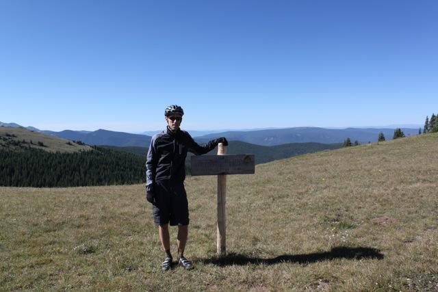 A person in cycling gear stands next to a wooden sign on a grassy hillside, with a panoramic view of distant mountains and a clear blue sky in the background. Monarch Crest Trail mountain bike trail.