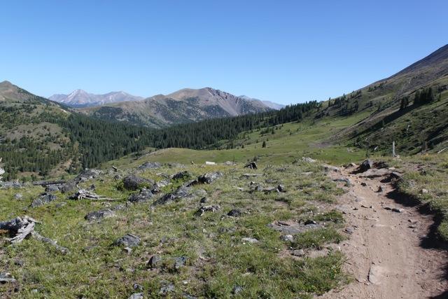 A scenic view of a mountain trail surrounded by lush green grass and rocky terrain, leading into a valley with forested hills and distant mountains under a clear blue sky. Monarch Crest Trail mountain bike trail.