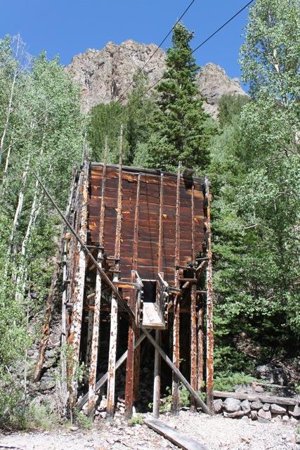 An old wooden mining structure surrounded by trees, with rocky mountains in the background and blue sky above. The building appears weathered and partially collapsed, supported by wooden beams. Monarch Crest Trail mountain bike trail.