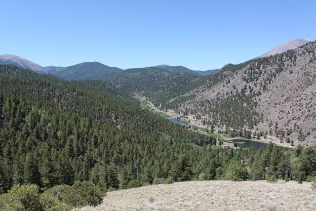 A scenic view of a mountainous landscape featuring dense forests, rolling hills, and a winding river below. The clear blue sky is visible above, with mountains in the distance. The foreground includes a mix of vegetation and open land. Monarch Crest Trail mountain bike trail.