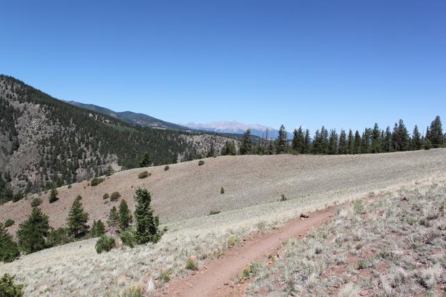 A panoramic view of a mountainous landscape featuring rolling hills, patches of greenery, and a dirt path winding through the terrain. In the background, the peaks of distant mountains are visible under a clear blue sky. Monarch Crest Trail mountain bike trail.