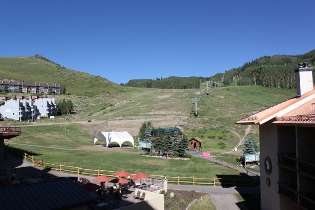 A scenic view of a mountainside with green hills under a clear blue sky. In the foreground, there are patio areas with outdoor seating, and a white event tent is visible on the grassy area. A ski lift ascends the slope, indicating a ski resort location. Buildings are partially visible on the left side, surrounded by trees. Evolution Bike Park at Crested Butte Mountain Resort mountain bike trail.