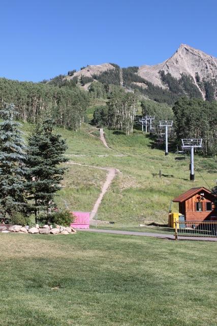 A scenic mountain landscape featuring a clear blue sky, lush green grass, and a winding dirt path leading up a hillside. In the foreground, there are coniferous trees and a small wooden building. Chairlift poles are visible in the background, indicating a ski resort or recreational area. Evolution Bike Park at Crested Butte Mountain Resort mountain bike trail.