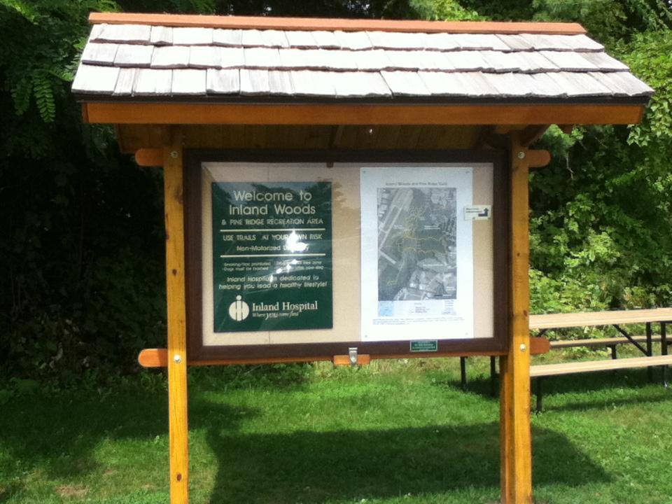Wooden information board in a park setting, displaying a welcome sign for Inland Woods and Pine Ridge Recreation Area, a map of trails, and safety instructions for trail use. The area features greenery and a bench in the background. Pine Ridge mountain bike trail.