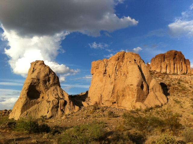 Rock formations rising from a desert landscape under a partly cloudy sky. The foreground features a tall, jagged rock on the left and a wider, more textured formation on the right, surrounded by sparse vegetation and sandy ground. Monolith Gardens mountain bike trail.